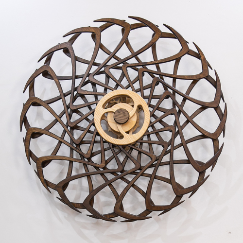 Dark-stained wooden wall sculpture with interlocking curved spokes radiating from a light central wheel, creating a mesmerizing kinetic pattern.