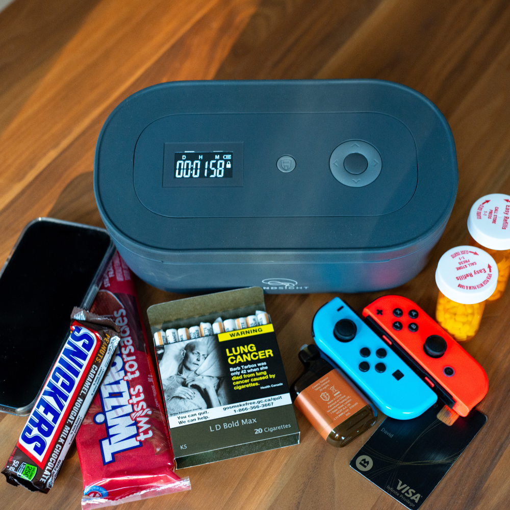A black Mindsight timed lockbox on a brown countertop with a phone, snacks, cigarettes, vape, controller, credit card and medication.