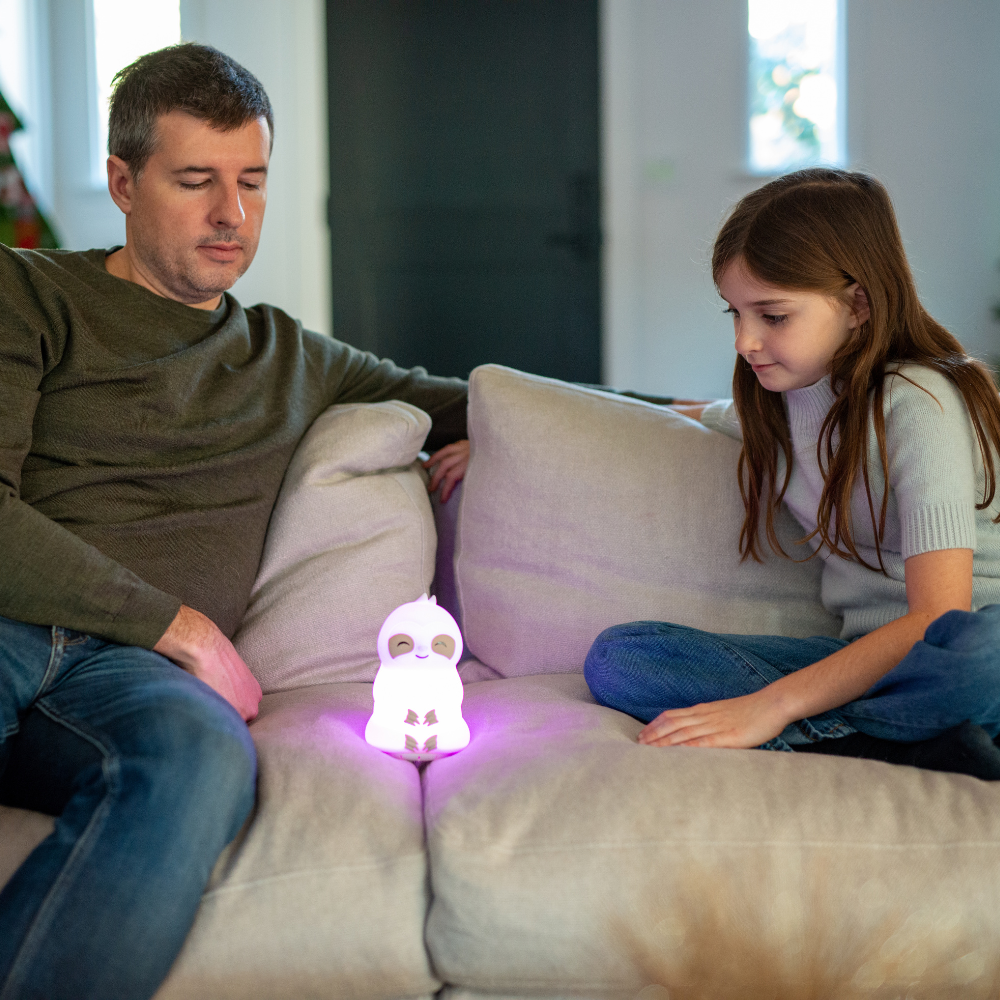 A father and his daughter sitting on a couch, meditating together while looking at a glowing owl-shaped device emitting soft purple light.