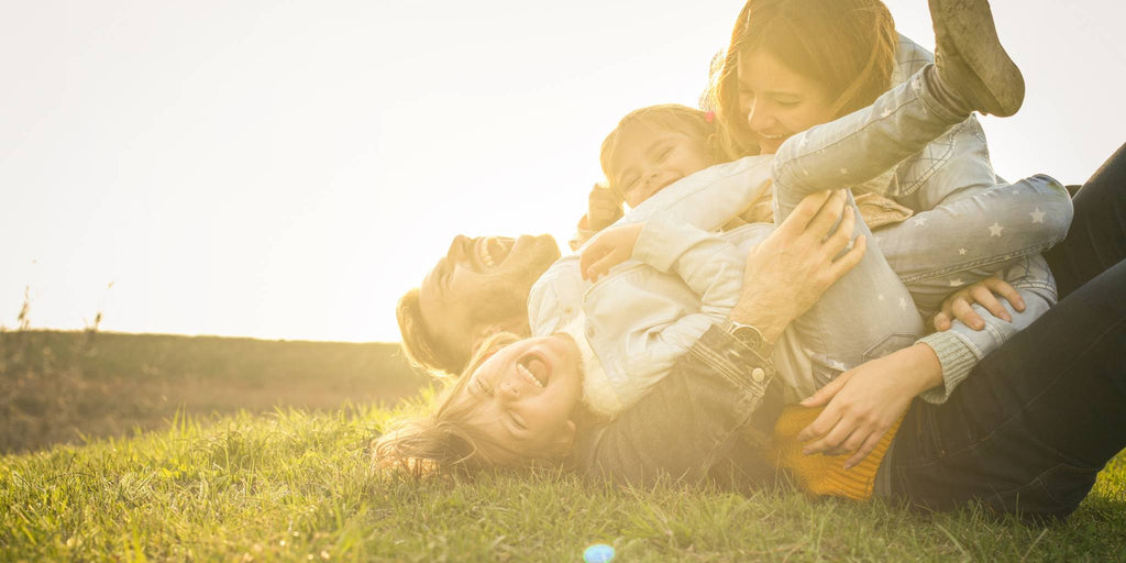 A family laughs and plays together on the grass, embracing in a joyful moment under the warm sunlight.